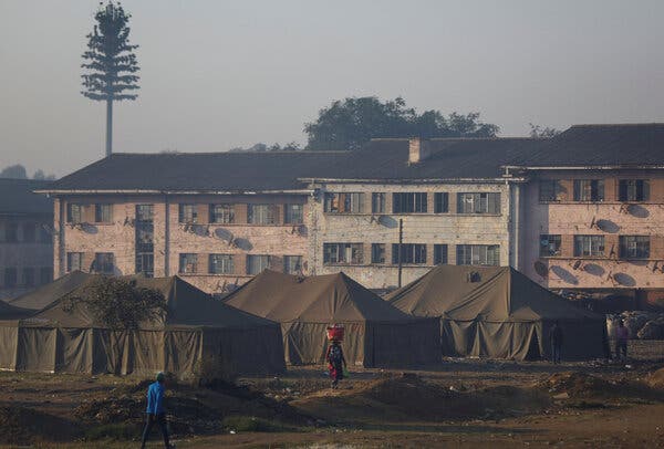 Three large olive-green tents set up in an open area in front of rundown buildings, some of which have window panes missing.