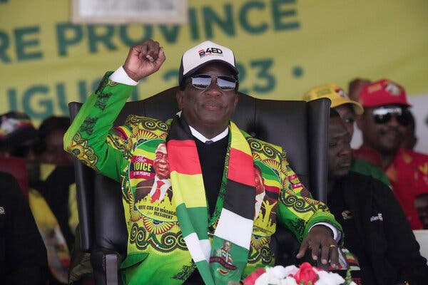 Emmerson Mnangagwa, wearing a baseball cap with the message “B4ED” and a yellow and green jacket with multicolor patterns and campaign slogans, sits on a black wingback chair and raises a fist.