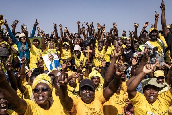 A crowd of people, mostly wearing yellow T-shirts, raise their arms. One person has a cross and another has a campaign poster for Nelson Chamisa.