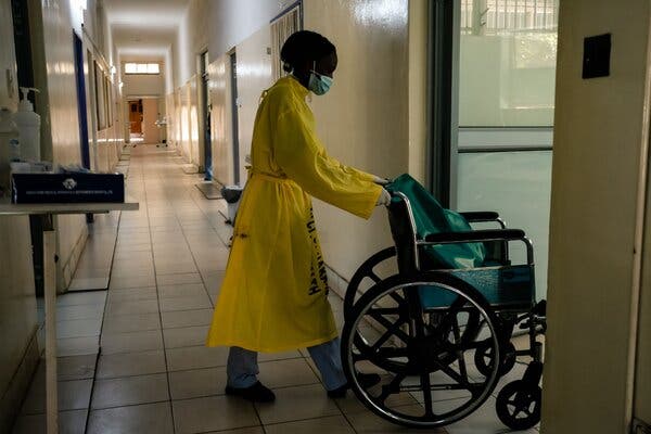 A health worker in a yellow gown pushes a wheelchair through a door in a hospital corridor.