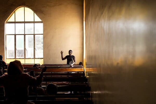 In a room with an arched window, a man with his arm raised and palm facing outward faces several rows of wooden pews with a few people seated in them 