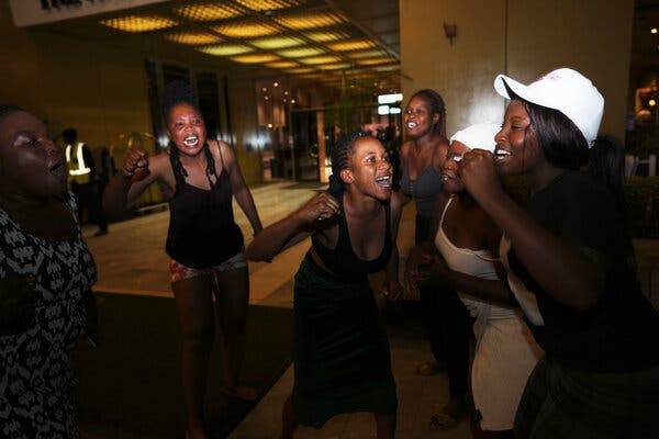 A group of laughing young women, one in a white cap, pump their fists in the hallway of a building with yellow light.