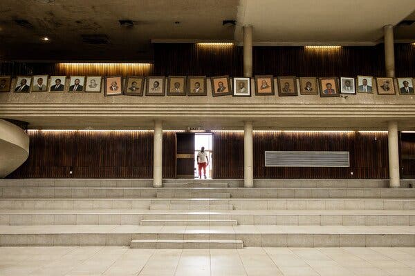 A person in a white top and red trousers stands in the doorway of a huge conference hall with cream columns and stairs. Above, on a balcony is a row of photos of ZANU-PF leaders.