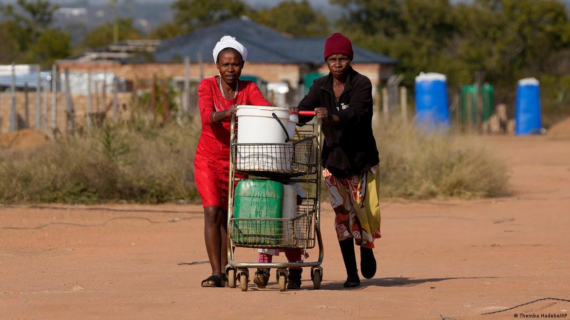 Two people pushing buckets filled with treated water on a shopping trolley along a dusty road