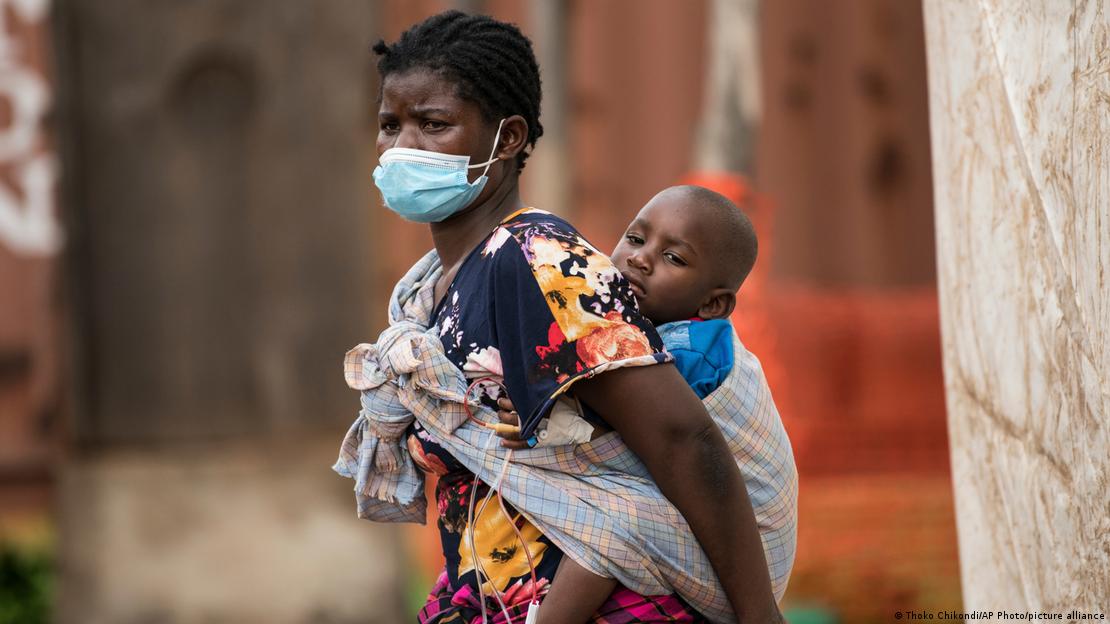 A woman wearing a face mask is carrying her ill son on her back